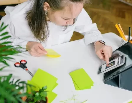 imagem de menina escrevendo em bloco de notas enquanto usa aplicativo de estudos no celular